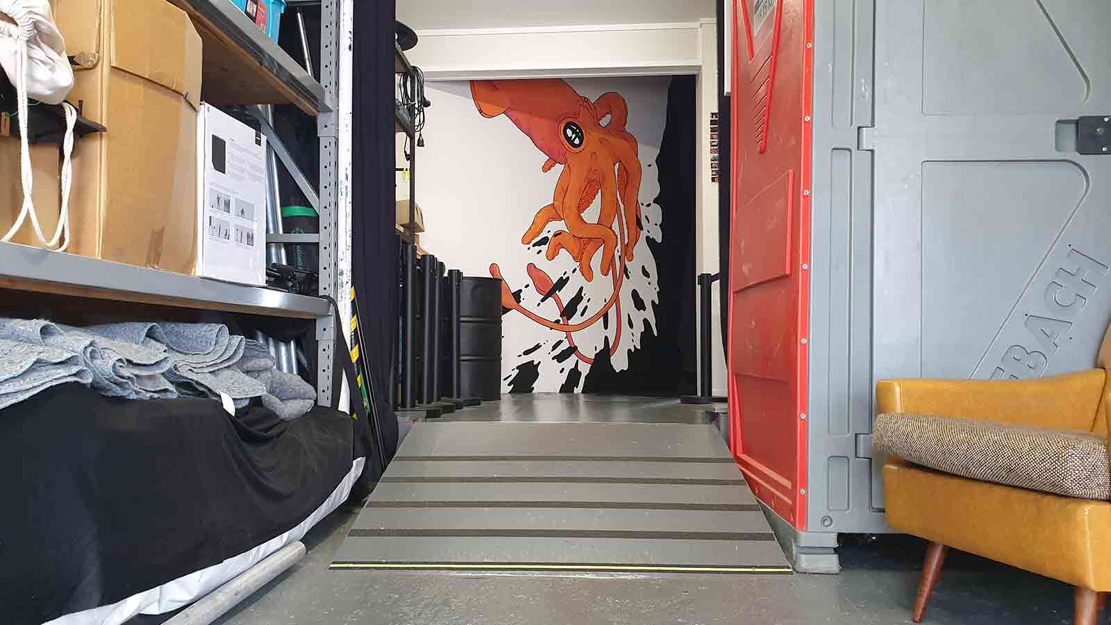 A corridor with storage shelfs either side leading toward an artistic image of a squid. Halfway along the corridoor is a ramp.